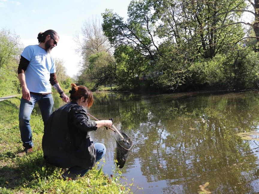 Two Subaru volunteers clean up a canal on a sunny spring day. One uses a net to remove debris from the water while the other stands nearby in support. Surrounded by greenery, the scene highlights Subaru's commitment to environmental stewardship. Impact