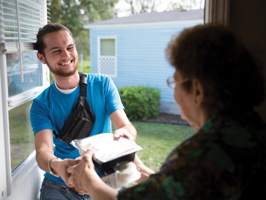 A smiling volunteer in a blue shirt hands a packaged meal to an older adult at her doorstep. Impact