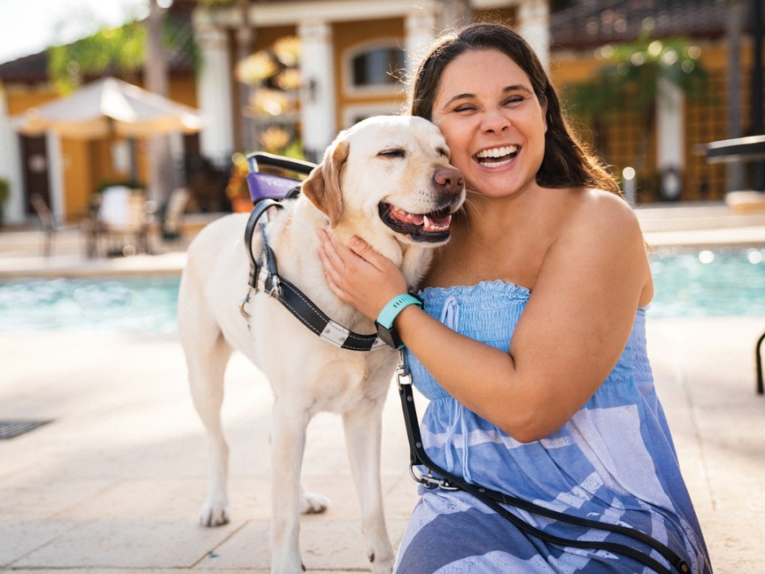 A visually impaired woman smiling while hugging her guide dog in an outdoor setting with a swimming pool in the background Impact