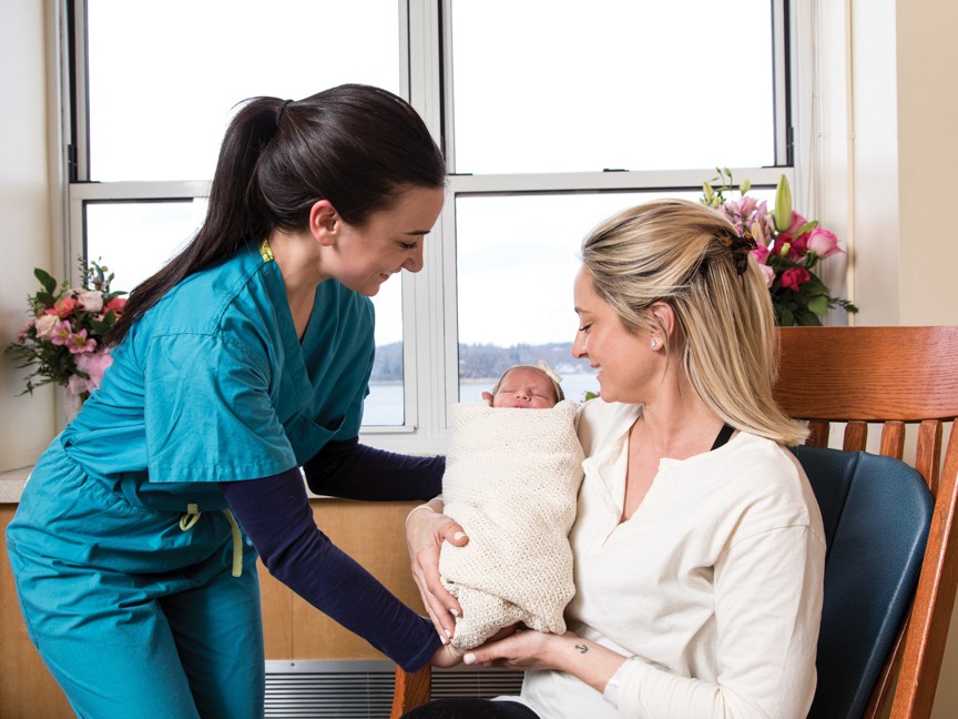 A mother sharing a proud moment with her new baby and their nurse after a healthy delivery. Impact